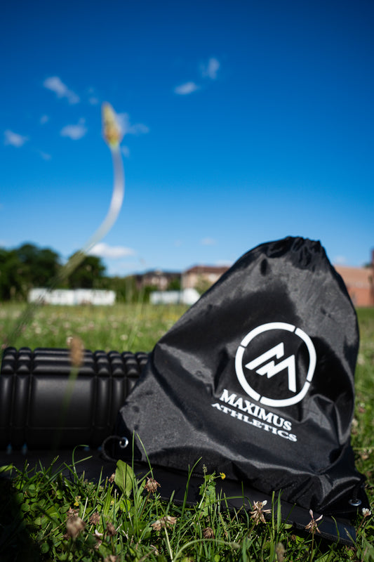 A black drawstring bag with the logo 'MAXIMUS ATHLETICS' on it, placed on a grassy field under a clear blue sky.